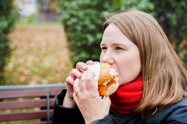 Mädchenfrau isst große HamburgerKäseburgerJunk fettiges leckeres StraßenessenTake-Away-BurgerMittagspause im Freien im Park auf der Bank sitzenUngesunde ErnährungWochenende entspannenKörper positivÜbergewichtsprobleme