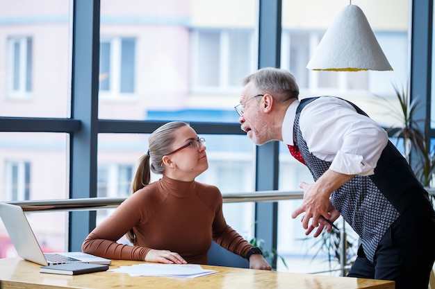 Mädchengeschäftsdame, die an einem Holztisch mit einem Laptop und einem Arbeitslehrer-Chefmentor sitzt, zeigt ihre Fehler an Schreit und schlägt mit der Hand auf den Tisch