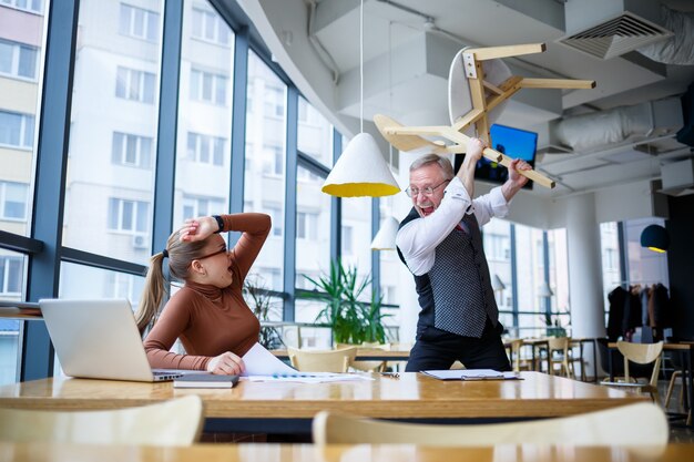 Mädchengeschäftsdame, die an einem Holztisch mit einem Laptop und einem Arbeitslehrer-Chefmentor sitzt, zeigt ihre Fehler an Schreit und schlägt mit der Hand auf den Tisch