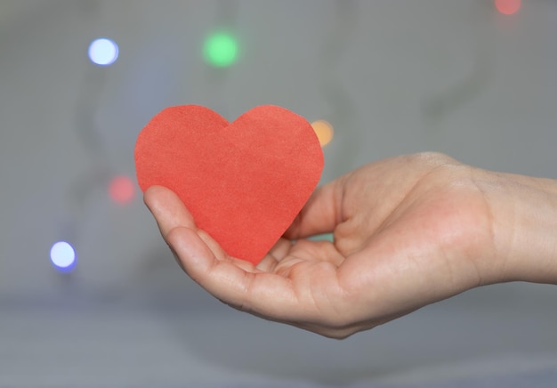 Mädchenhand, die rotes Papierherzweihnachtslichter im Hintergrund hält