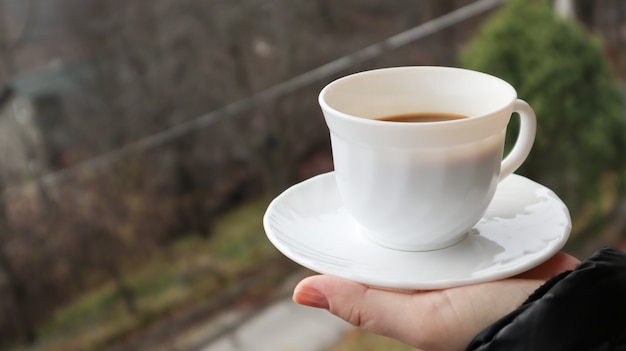 Mädchenhand hält eine Tasse weißen Kaffee und hat einen unscharfen grünen Naturhintergrund. Becher in einer weiblichen Hand auf einem Hintergrund von Bergen.