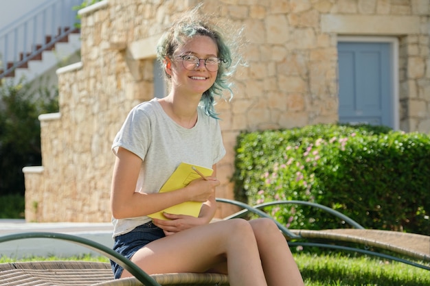 Mädchenjugendlicher, der mit Bleistiftnotizbuch studiert. Studentin sitzt im Gartenstuhl im Garten, sonniger Sommertag