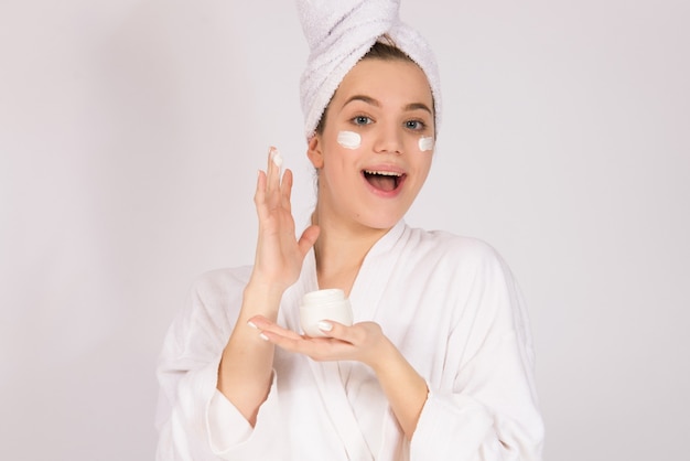 Mädchenmodell mit Gesichtscreme und Handtuch auf ihrem Kopf, Konzept der Hautalterungspflege und der Hautprodukte, auf weißem Hintergrund mit Kopienraum