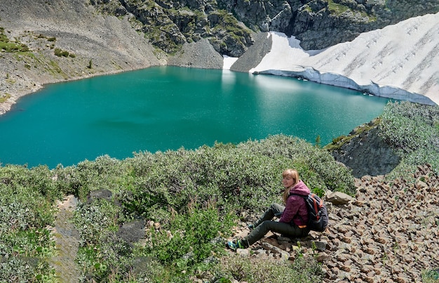 Mädchentouristin mit einem Rucksack sitzt auf den Felsen in den Bergen nahe dem blauen See. Altai Russland