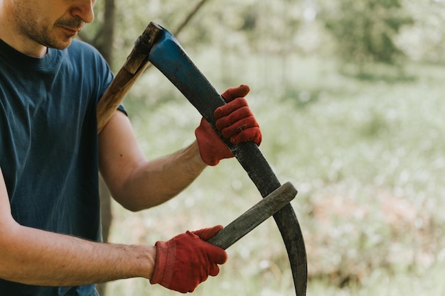 Mähen von Gras auf traditionelle altmodische Art und Weise mit Handsense auf dem Bauernhof des Haushaltsdorfs junger reifer Bauer, der die Sense mit Gras oder Wetzstein schärft, um das gewachsene Unkraut eines Ackerlandes zu mähen