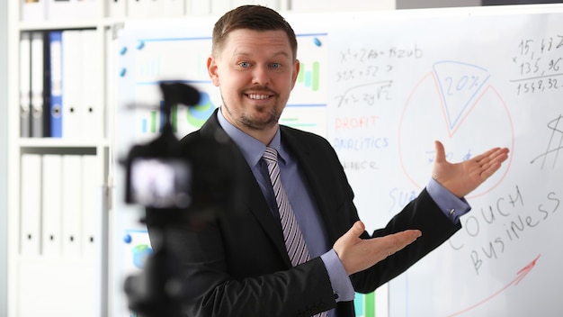 Männchen in Anzug und Krawatte zeigen Statistik-Grafikblock, der Promo-Videoblog oder Fotosession im Büro-Camcorder zur Stativnahaufnahme macht. Informationen zur Vlogger Selfie-Verkaufslösung oder zur Verwaltung von Finanzberatern