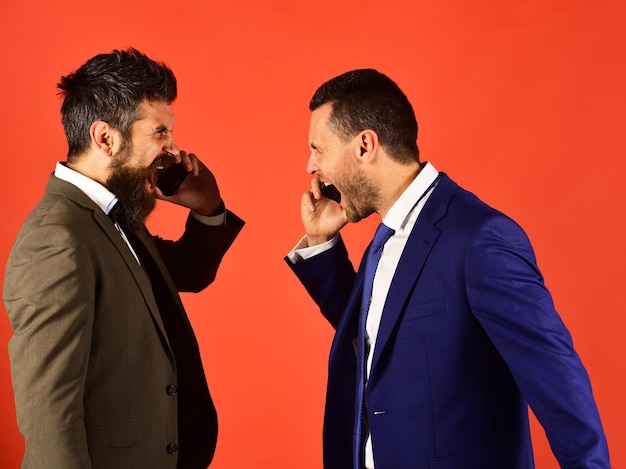 Männer mit Bärten stehen von Angesicht zu Angesicht auf rotem Hintergrund Geschäftsleute mit wütenden Gesichtern halten Mobiltelefone Business-Gesprächskonzept Machos in klassischen Anzügen schreien auf dem Handy