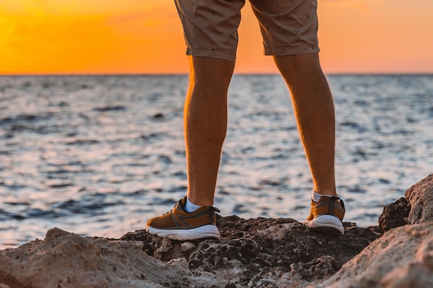 Foto männerbeine in shorts und turnschuhen stehen auf den felsen vor einem orangefarbenen sonnenuntergang am meer