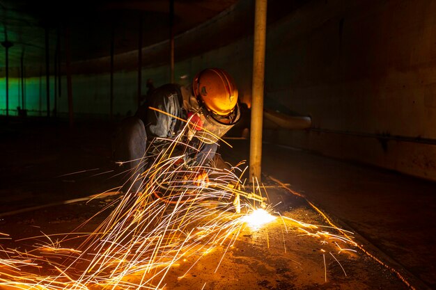 Männliche Arbeiter Metallschneidfunken auf der Stahlplatte des Tankbodens mit Blitzlicht, Nahaufnahme, Schutzhandschuhe und Maske in seitlich begrenztem Raum tragen.