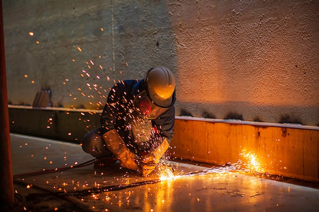 Männliche Arbeiter Metallschneidfunken auf der Stahlplatte des Tankbodens mit Blitzlicht, Nahaufnahme, Schutzhandschuhe und Maske in seitlich begrenztem Raum tragen.