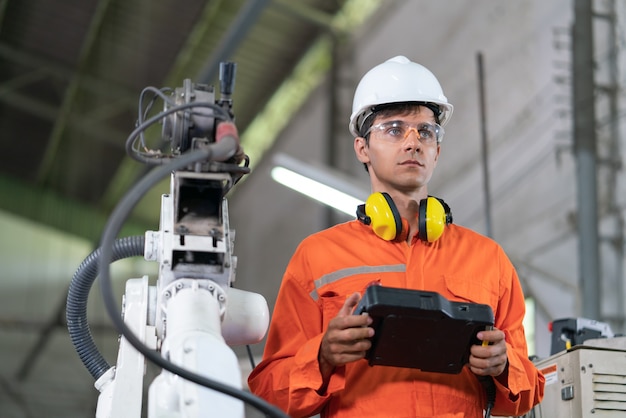 Männliche Automatisierungsingenieure tragen eine orangefarbene Uniform mit Helmsicherheit, die eine Roboterarmschweißmaschine mit einer Remote-Systemplatine in einer Industriefabrik steuert. Konzept der künstlichen Intelligenz.