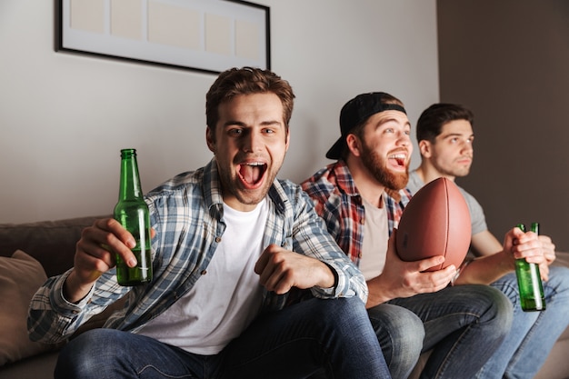 Männliche fröhliche Junggesellen, die schreien, während sie zu Hause feiern und Fußball mit Bier spielen sehen