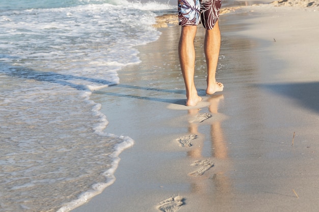 Männliche Füße am Ufer eines Sandstrandes an einem sonnigen Tag. junger Mann geht am Rand des Wassers entlang und hinterlässt Fußspuren. Platz für Text. Tourismus und Ruhe.