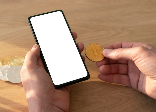 Männliche Hände, die Handy mit Bildschirm für Mock-up und Bitcoin-Münze in der Hand über Holztisch halten.