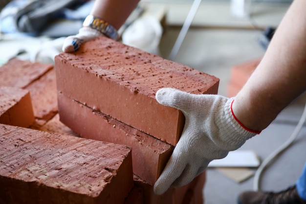 Männliche Hände, die mehrere rote Ziegelsteine auf Baustellennahaufnahme anheben