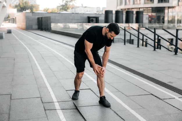 Männliche Hände, die schmerzendes Knie halten Starker Mann, der beim Joggen im Freien Schmerzen in seinem Fuß verspürt. Nahansicht. Männlicher Läufer in schwarzen Turnschuhen, der im linken Bein steht und Krämpfe verspürt. Schnittansicht.