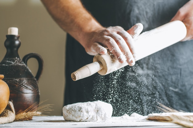 Männliche Hände gießen Mehl auf ein Nudelholz neben Teig, Tontopf, Ölflasche und Nudelholz auf dunklem Tisch, während sie kochen