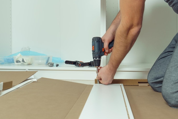 Männliche Hände halten schnurlose elektrische Schraubendreher, um die Wände der weißen Schublade zu befestigen, die die Kommode auf dem Boden zusammenbaut