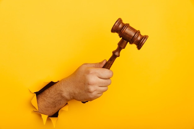 Männliche Hand, die den Hammer eines Richters durch zerrissenen gelben Hintergrund hält Gesetz und Auktion aconcept