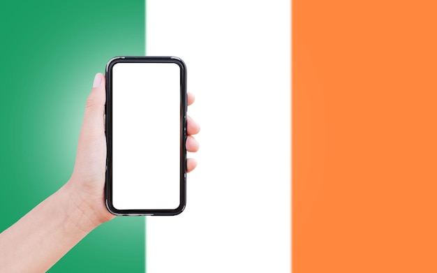 Männliche Hand, die ein Smartphone mit Leerzeichen auf dem Bildschirm auf dem Hintergrund der unscharfen Flagge Irlands hält Nahaufnahme