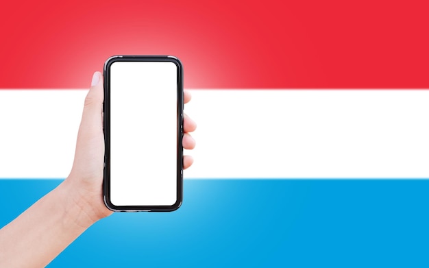 Männliche Hand, die ein Smartphone mit Leerzeichen auf dem Bildschirm auf dem Hintergrund der unscharfen Flagge Luxemburgs hält Nahaufnahme