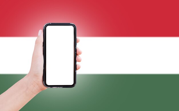 Männliche Hand, die ein Smartphone mit Leerzeichen auf dem Bildschirm auf dem Hintergrund der unscharfen Flagge Ungarns hält Nahaufnahmeansicht