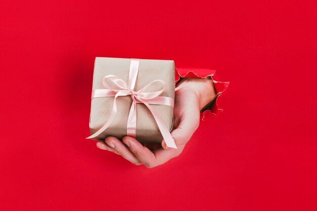 Männliche Hand, die Geschenkbox eingewickelt im Kraftpapier mit rosa Band durch ein Loch im Rot hält.