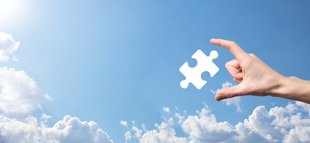 Männliche Hand, die Puzzlespielikone auf blauem Hintergrund hält. Stücke, die die Fusion von zwei Unternehmen oder Joint Venture, Partnerschaft, Fusionen und Übernahmekonzept darstellen.