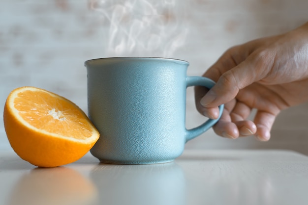 Männliche Hand, die Tasse mit heißem Getränk drinnen nimmt, Nahaufnahme. Dampf steigt aus der Tasse, Orangenscheibe auf dem Tisch. Frühstückskonzept, guten Morgen