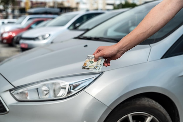 Männliche Hand, die viele Dollar gegen graues Auto im Rücken, Verkauf oder Miete hält