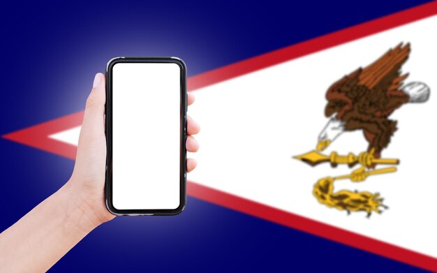 Männliche Hand hält Smartphone mit leerem Bildschirm auf dem Hintergrund der unscharfen Flagge von Amerikanisch-Samoa Nahaufnahme