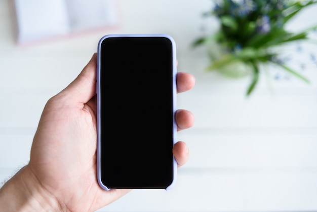 Männliche Hand mit einem Smartphone. Schwarzer leerer Bildschirm. Tabelle mit Notizbuch und Blumen ein