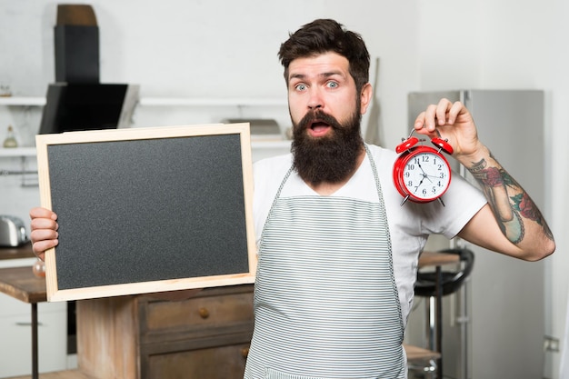 Männliche Haushälterin in der Küche, die nach Rezept bärtige Hipster kocht, bietet Menürestaurantpersonal, Kopienraum, überraschter Mann mit Tafel und Weckerzeit für gesunde Speisen und Getränke