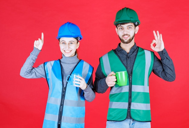 Männliche und weibliche ingenieure mit helm, der blaue und grüne becher hält und zufriedenheitszeichen zeigt.