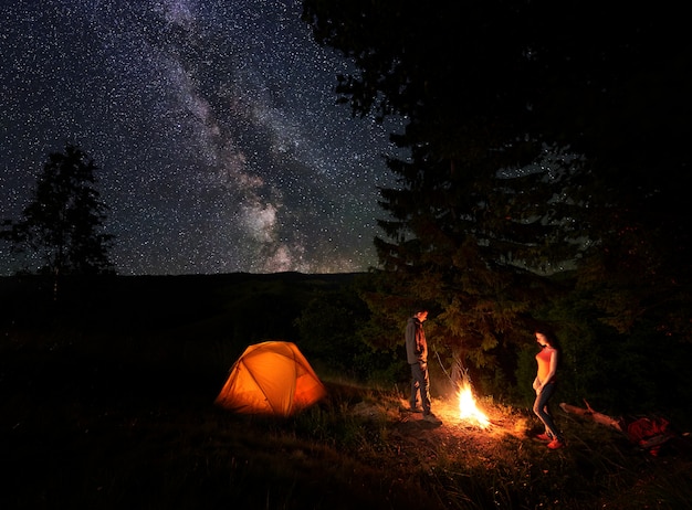 Männliche und weibliche Wanderer ruhen sich am Lagerfeuer in der Nähe des Waldes aus und leuchten in einem orangefarbenen Zelt beim Campen in den Bergen unter dem unglaublich schönen Sternenhimmel, der deutlich in der Milchstraße zu sehen ist.