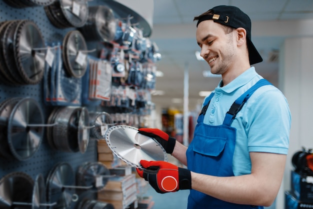 Männlicher Arbeiter in Uniform, der kantige Scheibe für Säge im Werkzeugspeicher wählt. Auswahl an professioneller Ausrüstung im Baumarkt, Instrumentensupermarkt