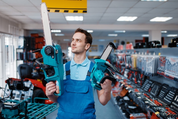 Männlicher Arbeiter in Uniform hält große und kleine Kettensägen im Werkzeugladen. Auswahl an professioneller Ausrüstung im Baumarkt, Instrumentensupermarkt