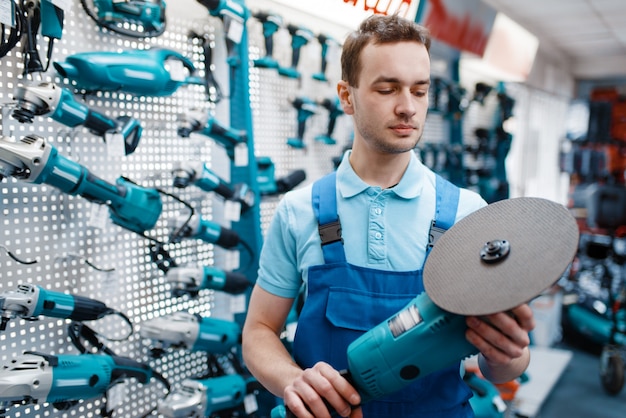 Männlicher Arbeiter in Uniform hält Winkelschleifer im Werkzeugspeicher. Auswahl an professioneller Ausrüstung im Baumarkt, Instrumentensupermarkt