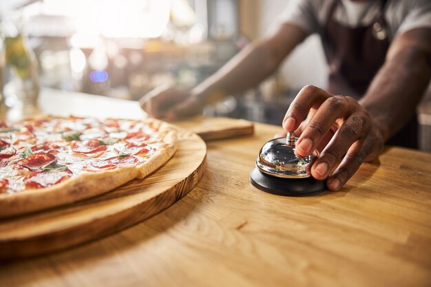 männlicher Arbeiter mit Rufglocke, während er mit frisch gebackener Peperoni-Pizza an der Theke steht?