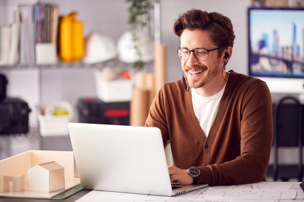 Männlicher Architekt im Büro, der an einem Plan am Schreibtisch arbeitet und einen Anruf über drahtlose Kopfhörer entgegennimmt