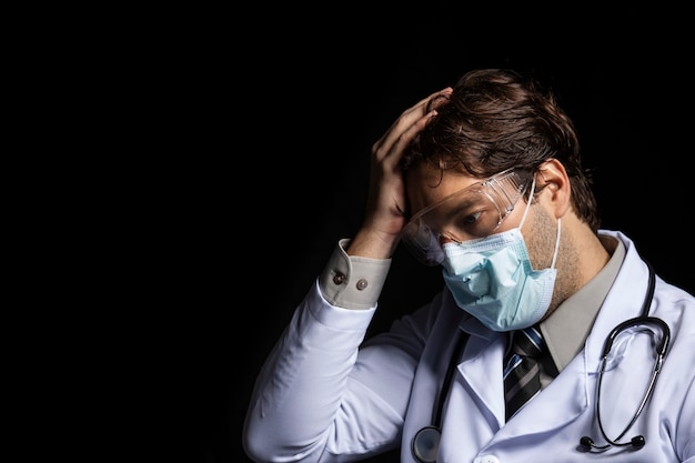 Männlicher Arzt, der eine Maske und eine Schutzbrille trägt, müde von der Arbeit mit covid-19 auf einem schwarzen Hintergrund.
