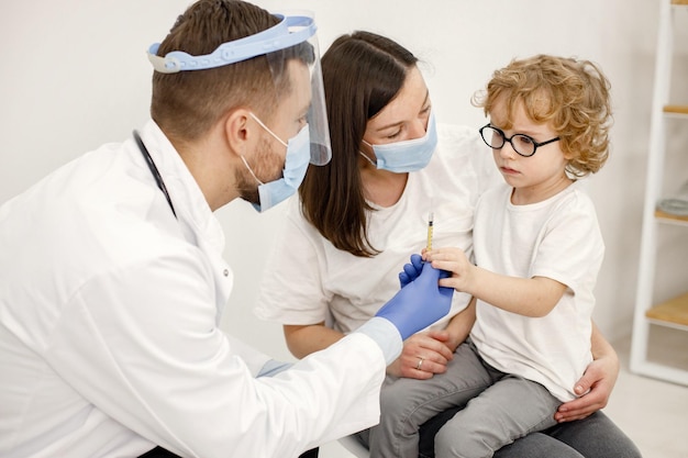 Männlicher Arzt, der einem kleinen Jungen, der auf dem Schoß der Mutter sitzt, eine Impfung vornimmt