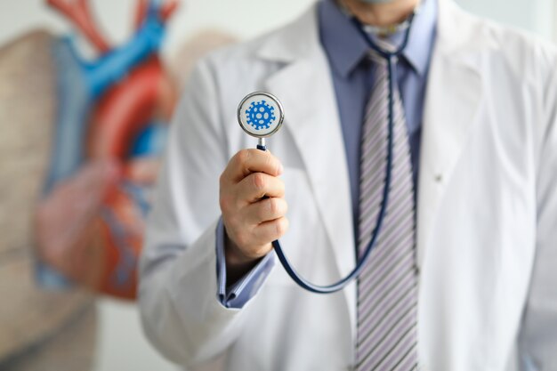 Männlicher Arzt hält in der Hand Stethoskop mit Coronavirus Symbol Nahaufnahme.