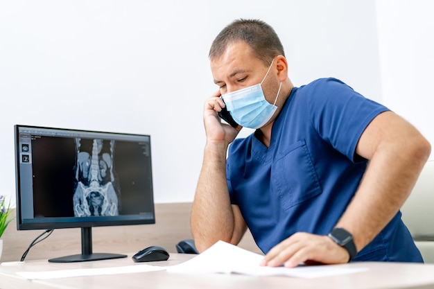 Männlicher Arzt im Büro, der am Computer arbeitet. Moderner Krankenhausbürohintergrund. Sprechen am Telefon.