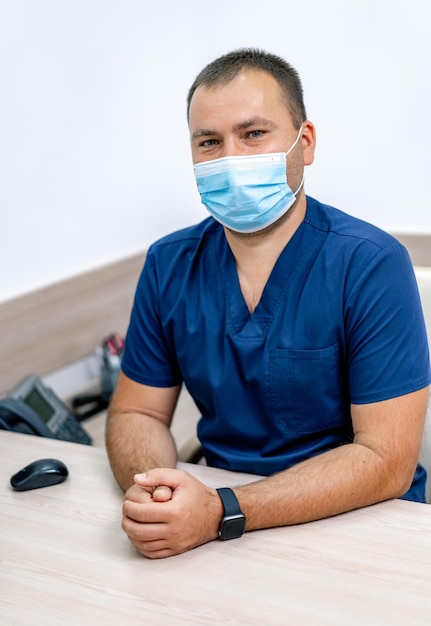 Männlicher Arzt im Büro mit Blick in die Kamera. Moderner Krankenhausbürohintergrund. Medizinische Maske im Gesicht. Nahaufnahme.