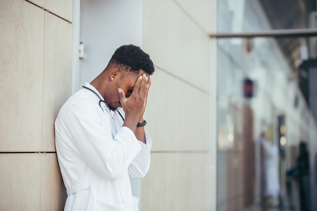 Männlicher Arzt in der Klinik, der seinen Kopf hält, hat gesundheitliche Probleme und ist nach einem harten Tag müde, depressiv hat nicht das gewünschte Ergebnis erzielt, afroamerikanischer Arzt ist traurig