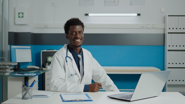 Männlicher Arzt in medizinischer Uniform sitzt am Schreibtisch mit modernem Laptop, Dokumentendateien und Gesundheitsgeräten im Büro der medizinischen Klinik. Professioneller Sanitäter, der die Kamera anschaut