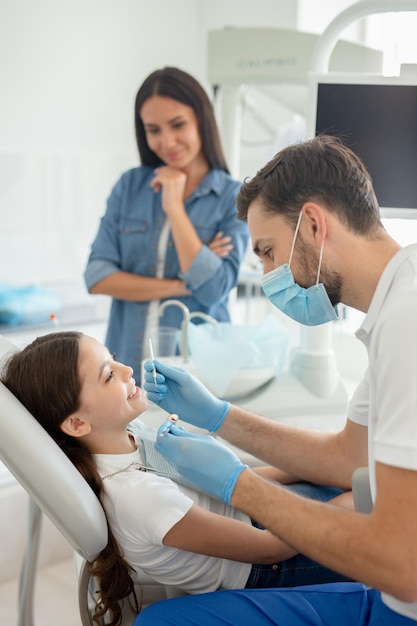 Männlicher Arzt mit Instrumenten in den Händen sieht Mädchenzähne aus, die im Behandlungsstuhl sitzen, während die Mutter neben ihr steht, um Unterstützung zu erhalten