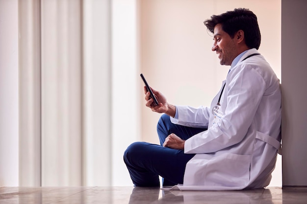 Männlicher Arzt mit weißem Kittel sitzt auf dem Boden im Krankenhauskorridor Textnachrichten auf dem Handy