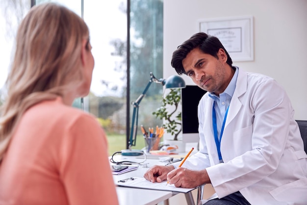 Männlicher Arzt oder Hausarzt, der sich mit einer reifen Patientin für einen Termin im Büro trifft und sich Notizen über die Zwischenablage macht